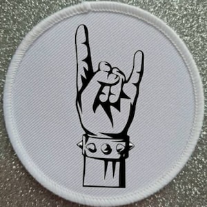 Może przedstawiać: Biała okrągła łatka z czarno-białym rysunkiem dłoni wykonującej gest rock and roll. Dłoń ma na sobie bransoletkę z ćwiekami.