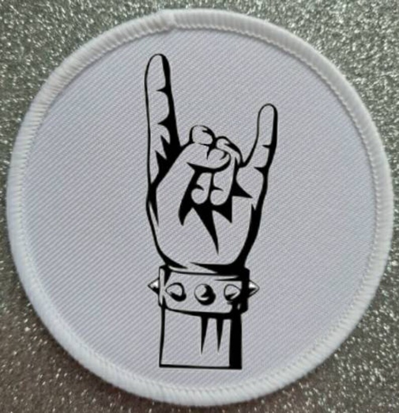 Pode incluir: Um remendo redondo branco com uma ilustra&ccedil;&atilde;o em preto e branco de uma m&atilde;o fazendo o gesto de rock and roll. A m&atilde;o est&aacute; usando uma pulseira com tachas.