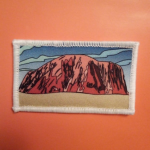 Può includere: Una toppa ricamata che raffigura una catena montuosa rossa e marrone contro un cielo blu con nuvole bianche. La toppa ha un bordo bianco con un bordo cucito.