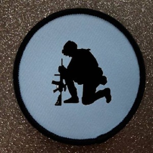 May include: A black and white embroidered patch of a soldier kneeling with a rifle. The patch is circular and has a light blue background.