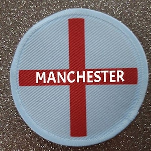 May include: A white and red circular patch with a red cross and the word "MANCHESTER" in white letters.