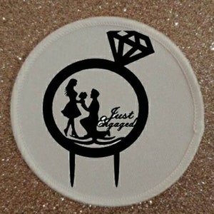 Puede incluir: Un adorno para pastel circular blanco con una silueta negra de una pareja y un diseño de anillo de diamantes. El texto "Just Engaged" también es negro. El adorno tiene dos púas para insertar.