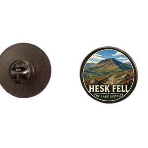 May include: A circular metal pin featuring a landscape illustration of a mountain with a path leading toward it. The text HESK FELL and THE LAKE DISTRICT is printed in white on a dark background. The back shows a metal clutch fastener mechanism.