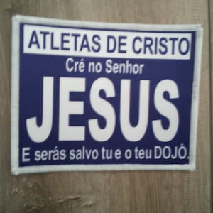 Peut inclure: Un patch bleu et blanc avec le texte "ATLETAS DE CRISTO" et "Crê no Senhor JESUS" en lettres blanches. Le texte "E serás salvo tu e o teu DOJO" est également en lettres blanches.