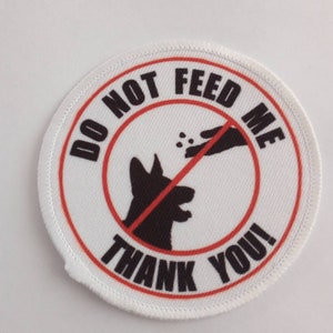 Puede incluir: Parche redondo blanco con texto rojo y negro y una línea roja que atraviesa una mano con una silueta negra de la cabeza de un perro. El texto dice "No me alimentes ¡Gracias!"