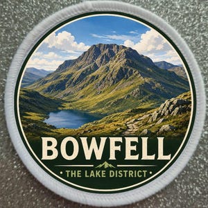 May include: A circular embroidered patch featuring a landscape of Bowfell mountain in the Lake District. The image shows a blue lake, green hills, and a rocky path under a blue sky with white clouds. The text reads BOWFELL THE LAKE DISTRICT.