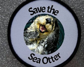 Può includere: Una toppa rotonda bianca e nera con una lontra marina sorridente al centro. Il testo "Save the Sea Otter" è scritto intorno alla lontra.