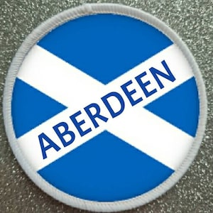 May include: A white and blue round patch with the Scottish flag and the word "ABERDEEN" in blue letters.