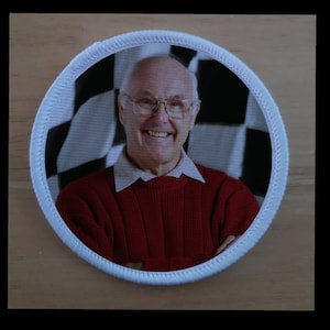 May include: A white circular patch with a red stitched border. The patch features a photo of a man smiling with a chequered background. He is wearing a red jumper and glasses.