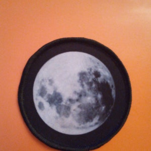 May include: A black and white moon patch with a detailed image of the moon's surface. The patch is circular and has a black border.