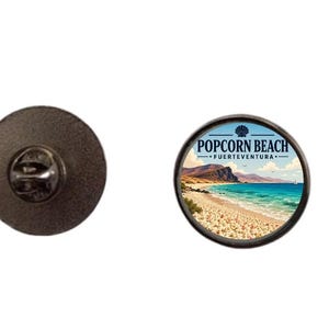 May include: A round metal pin badge featuring a scenic illustration of Popcorn Beach in Fuerteventura. The design displays a sandy shore with white pebbles, turquoise ocean water, and distant cliffs. The text reads Popcorn Beach Fuerteventura.