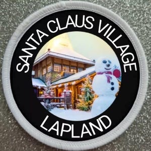 May include: A white round patch with black text and a photo of a snowy village with a snowman in the foreground. The text reads "Santa Claus Village Lapland".