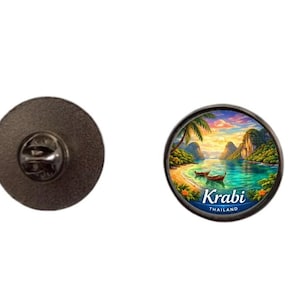 Puede incluir: Pin redondo con una imagen escénica de Krabi, Tailandia, con una playa, barcos y montañas bajo un cielo de atardecer. El pin tiene un respaldo de metal negro.