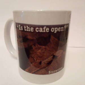 Może przedstawiać: Biały kubek ceramiczny z brązowym i czarnym obrazem pasma górskiego i tekstem "Is the cafe open?" oraz "Snowdon" nadrukowanym na nim.