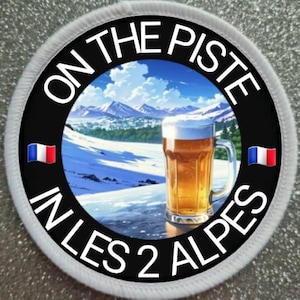 Può includere: Un cerotto rotondo bianco e nero con il testo "On the Piste in Les 2 Alpes" e un'immagine di una scena di montagna innevata con un bicchiere di birra in primo piano. Il cerotto ha due piccole bandiere francesi su entrambi i lati.