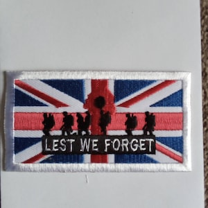 以下が含まれることがあります： 赤、白、青のユニオンジャックの旗のデザインが施された白い刺繍パッチ。兵士のシルエットが旗の上を歩き、中央にはポピーが描かれている。旗の下には「LEST WE FORGET」の文字が書かれている。