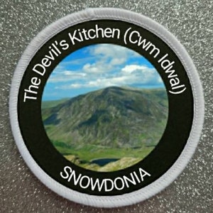 Puede incluir: Un parche redondo blanco con texto negro y un círculo negro en el centro. El texto dice "The Devil's Kitchen (Cwm Idwal) SNOWDONIA". El círculo contiene una foto de una cordillera.