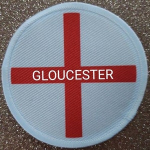 Könnte beinhalten: Ein weißer runder Patch mit einem roten Kreuz und dem Wort "GLOUCESTER" in der Mitte.