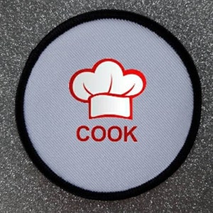 Könnte beinhalten: Runder Stoff-Patch in Weiß mit schwarzem Rand. Der Patch zeigt eine rote Kochmütze mit weißem Rand und das Wort "COOK" in roten Buchstaben.