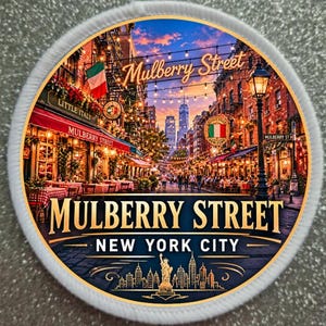 May include: A round patch featuring a detailed illustration of Mulberry Street in New York City. The image showcases a bustling street scene with restaurants, string lights, and the text "Mulberry Street" and "New York City".