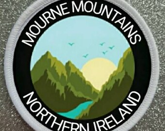 Peut inclure: Écusson rond blanc avec du texte noir et un graphique d'une chaîne de montagnes avec une rivière bleue et un soleil jaune. Le texte indique "Mourne Mountains Northern Ireland".