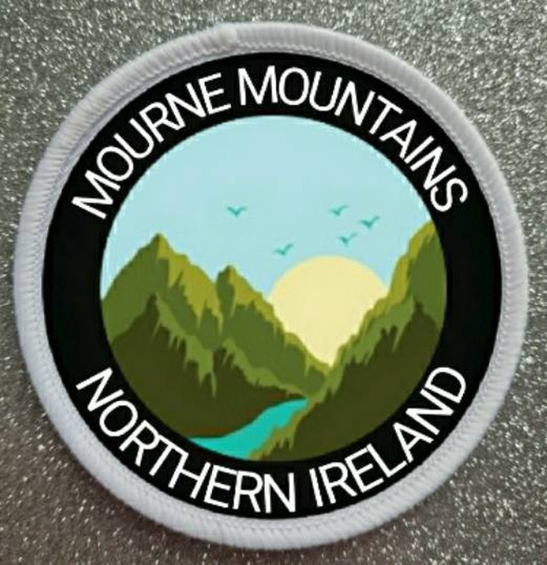 May include: White round patch with black text and a graphic of a mountain range with a blue river and a yellow sun. The text reads "Mourne Mountains Northern Ireland".