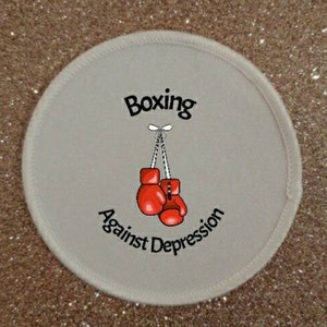 Peut inclure: Un patch rond blanc avec le texte "Boxing Against Depression" et une image de dessin animé de gants de boxe rouges suspendus à une ficelle.