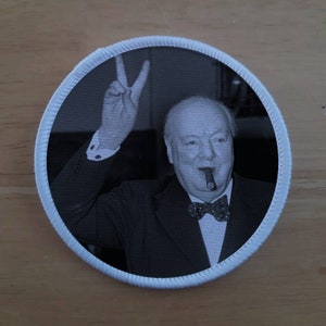May include: A black and white photo of Winston Churchill giving a peace sign while holding a cigar. The photo is framed in a white circle.