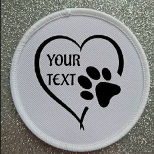 Puede incluir: Parche redondo de tela blanca con una huella de pata negra dentro de un corazón. El texto "YOUR TEXT" está dentro del corazón.