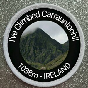 May include: Circular patch with the text "I've Climbed Carrauntoohil" in white, encircling a mountain image. The patch also includes "1038m - IRELAND". The patch has a black center, white border, and is set against a glittery background.