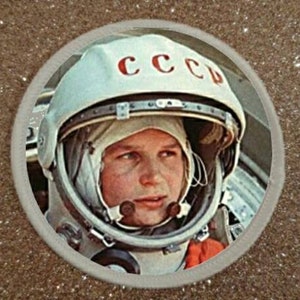 Può includere: Un primo piano di un casco da astronauta bianco con la scritta "CCCP" stampata sul davanti. L'astronauta indossa una tuta spaziale bianca con una striscia rossa sulla spalla.