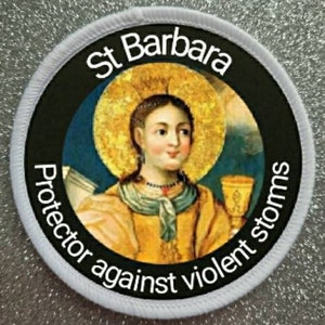 May include: A circular patch depicting St Barbara with a golden halo and yellow robes. The text "St Barbara" is at the top, with "Protector against violent storms" around the edge. The patch has a white stitched border.