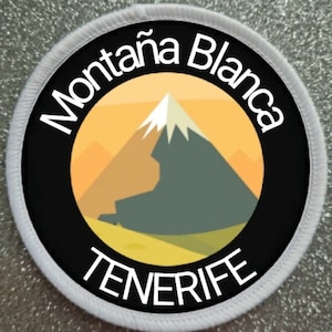 Puede incluir: Parche redondo blanco con borde negro y texto blanco. El texto dice "Montaña Blanca TENERIFE". Dentro del borde hay una montaña amarilla y naranja con un pico blanco.