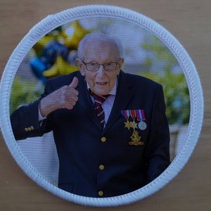 Puede incluir: Un parche redondo blanco con una foto de un hombre mayor con un traje azul oscuro y una corbata a rayas rojas y blancas. Está haciendo un pulgar hacia arriba y sonriendo. Lleva varias medallas en su chaqueta.