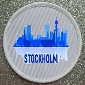 May include: White oval fabric patch featuring a blue silhouette of the Stockholm skyline and the word "STOCKHOLM" in blue text.