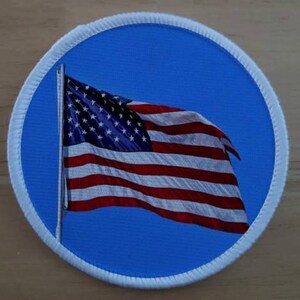 May include: A white circle patch with a blue background and a red, white, and blue American flag waving in the wind.