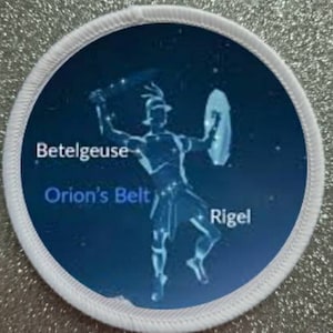 May include: A white circular patch with a blue background featuring a constellation of stars in the shape of a person. The constellation is labeled "Betelgeuse", "Orion's Belt", and "Rigel".