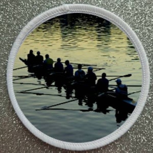 May include: A group of people rowing a boat on a lake at sunset. The water is calm and the sky is a soft orange and yellow. The people are silhouetted against the sky.