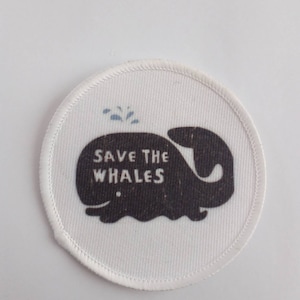 May include: Circular white patch with a stitched edge. It displays a dark grey whale illustration and the white text "SAVE THE WHALES". Blue water spouts are depicted above the whale.