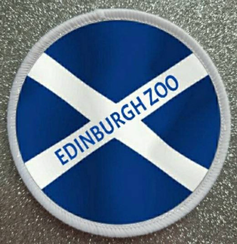 Puede incluir: Un parche redondo blanco y azul con el texto "EDINBURGH ZOO" en letras blancas sobre un fondo azul. El parche tiene un borde blanco.