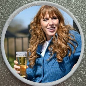 Peut inclure: Un écusson rond avec la photo d'une femme tenant un verre de bière. La femme a de longs cheveux roux bouclés et porte une veste bleue sur une chemise blanche. L'écusson a une bordure blanche et est posé sur un fond argenté scintillant.