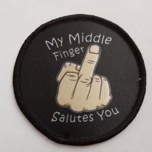 Può includere: Un cerotto rotondo nero con testo bianco che recita "My Middle Finger Salutes You". Una mano di cartone animato con il dito medio alzato è al centro del cerotto.