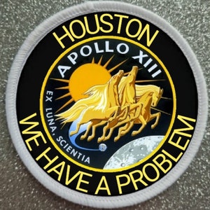 Pode incluir: Um remendo redondo preto e branco com uma borda branca. O remendo apresenta um sol amarelo, uma lua e três cavalos dourados correndo sobre um fundo preto. O texto "HOUSTON" está em amarelo na parte superior do remendo, e "APOLLO XIII" está em amarelo abaixo do sol. O texto "WE HAVE A PROBLEM" está em amarelo ao redor da parte inferior do remendo. O texto "EX LUNA, SCIENTIA" está em branco abaixo dos cavalos.