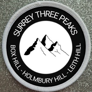 Pode incluir: Etiqueta redonda branca e preta com o texto "SURREY THREE PEAKS" e "BOX HILL - HOLMBURY HILL - LEITH HILL" em torno de uma ilustração em preto e branco de três montanhas.
