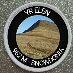 May include: White round patch with a black border and white text. The text reads "Yr Elen 962 M - Snowdonia". The patch features a photo of a brown hill with a blue sky in the background.