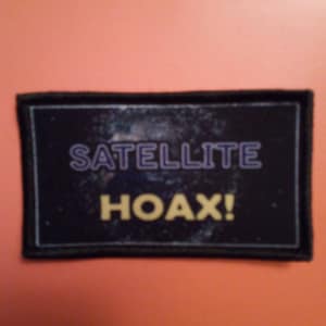 May include: A black patch with white stitching around the edges. The patch has a black background with a galaxy-like image and the text "SATELLITE HOAX!" in yellow.