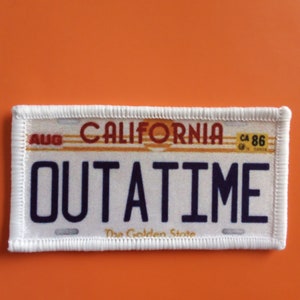 May include: White California license plate patch with blue text that reads "OUTATIME" and the words "The Golden State" at the bottom. The license plate number is CA 86.