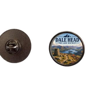 Könnte beinhalten: Ein runder Ansteck-Pin aus Metall mit einer Landschaftsaufnahme von Dale Head im Lake District. Das Motiv zeigt einen Steinhaufen auf einem grasbewachsenen Bergpfad mit Blick auf einen blauen See und Hügel, mit dem Text DALE HEAD THE LAKE DISTRICT.
