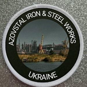 May include: A white circular patch with black text that reads "Azovstal Iron & Steel Works Ukraine". The patch features a black and white photo of a steel mill.