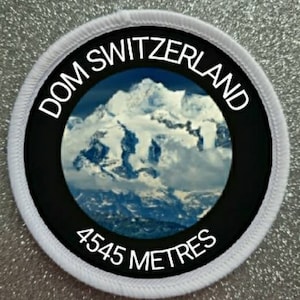 Könnte beinhalten: Runder weißer Patch mit schwarzem Rand und weißem Text. Der Text lautet "DOM SWITZERLAND 4545 METRES". Die Mitte des Patches zeigt ein Foto eines schneebedeckten Berggipfels.